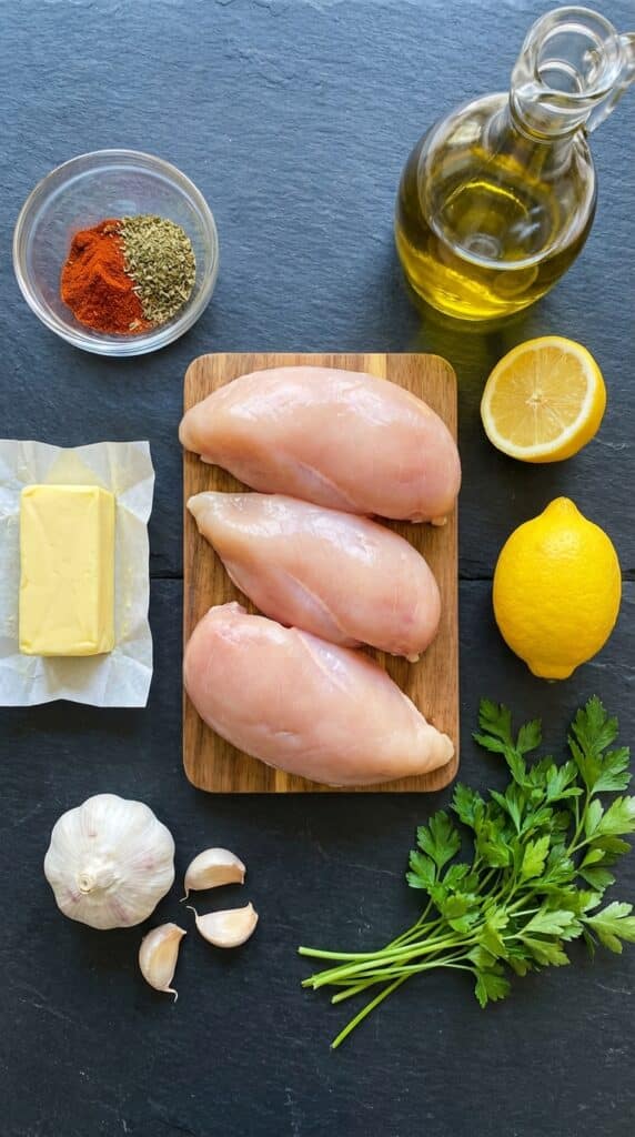 A flat lay showing raw chicken breasts, butter, garlic cloves, fresh lemon, olive oil, parsley, and dry spices on a dark slate board.