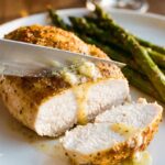 A close-up of a knife slicing into a juicy baked chicken breast, revealing the moist white interior, with garlic butter dripping down the side.