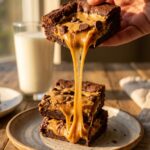A close-up of a hand lifting a thick chocolate bar filled with peanut butter chips, creating a long, gooey caramel stretch.