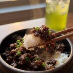 A close-up of chopsticks lifting a bite of glazed Korean ground beef and white rice from a bowl.