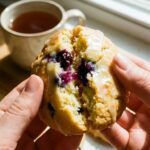 A close-up of hands pulling apart a glazed lemon blueberry cookie, revealing a soft center and a burst juicy blueberry.