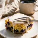 A close-up of a fork cutting into a thick slice of blueberry cake with lemon glaze on a plate, with coffee in the background.