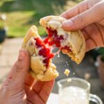 Two hands breaking a soft lemon raspberry cookie in half, showing a juicy red raspberry pocket and melted white chocolate inside.