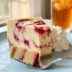 A close-up of a slice of lemon raspberry swirl cheesecake showing the red raspberry ribbons inside the yellow filling, with a fork on a pastel plate.