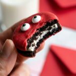 A close-up of a hand holding a decorated love bug Oreo cookie with a bite taken out, showing the chocolate cookie and cream inside.