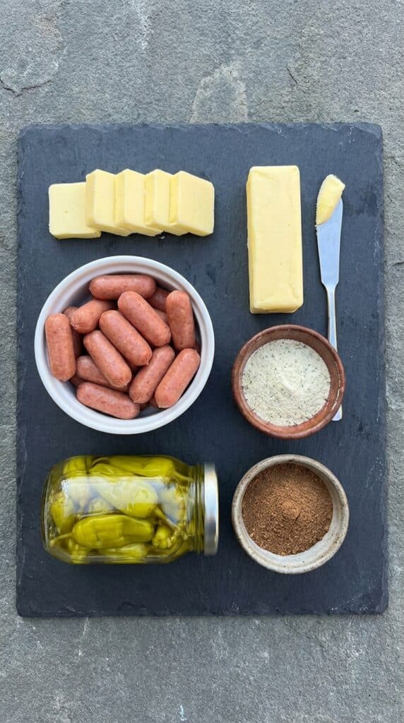 A flat lay showing mini sausages, butter slices, a jar of pepperoncinis, ranch seasoning, and au jus powder on a dark slate board.