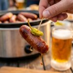 A close-up of a hand holding a toothpick with a glazed mini sausage and a pepperoncini, with a beer in the background
