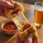 A close-up of hands pulling apart a hot pepperoni stuffed croissant roll, showing an extreme melted mozzarella cheese stretch.