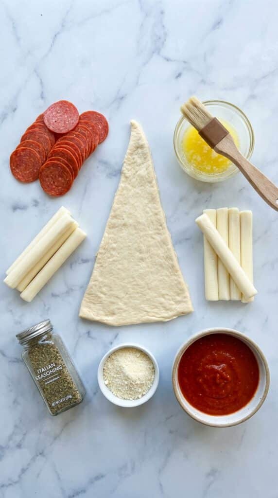A flat lay showing a raw crescent dough triangle, pepperoni slices, mozzarella sticks, melted butter, spices, and marinara sauce on a marble counter.