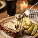 A close-up of a fork cutting into a thick slice of cheesy mushroom meatloaf, served with mashed potatoes and dill pickles.