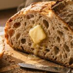 A close-up of a thick slice of crusty artisan bread with large air pockets, topped with melting salted butter.