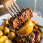 A close-up of a fork holding a glistening piece of beef and a baby potato covered in garlic butter, with a plate and red wine in the background.