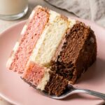 A close-up of a fork cutting through a thick slice of three-layer Neapolitan cake on a pink plate, showing the moist cake crumb.