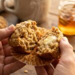 A close-up of hands pulling apart a warm oatmeal muffin, with steam rising and butter melting into the soft interior.