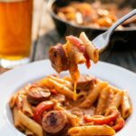 A close-up of a fork lifting a bite of penne pasta and smoked sausage coated in a thick, creamy cajun sauce.