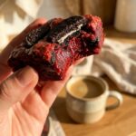 A close-up of a hand holding a half-eaten red velvet brownie, showing a dense fudgy texture and a baked Oreo inside.