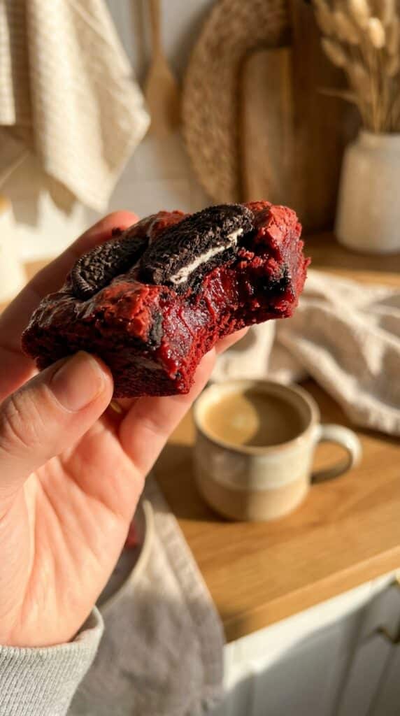 A close-up of a hand holding a half-eaten red velvet brownie, showing a dense fudgy texture and a baked Oreo inside.