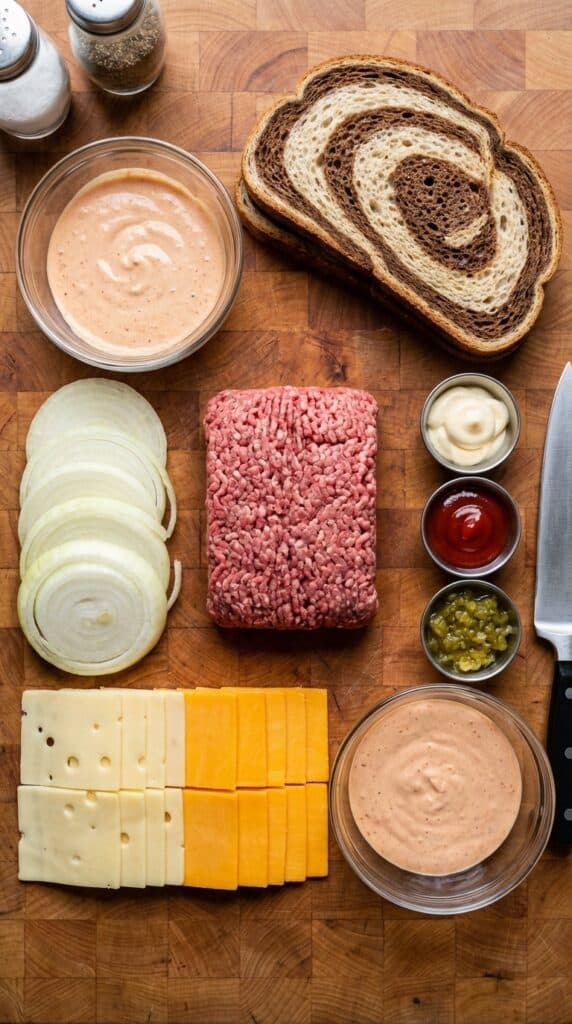 A flat lay showing raw ground beef, rye bread, sliced onions, cheese slices, and ingredients for secret sauce on a wooden board.