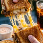 Hands pulling apart a hot patty melt sandwich, creating a long, gooey cheese stretch with steam rising from the beef and onions.