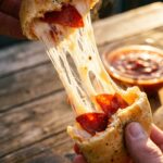 A close-up of hands pulling apart a baked pizza bomb, revealing an extreme melted mozzarella cheese pull and pepperoni inside.