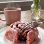 A close-up of a fork pulling apart a warm pink cinnamon roll, showing the fluffy pink interior and dark cinnamon swirl.