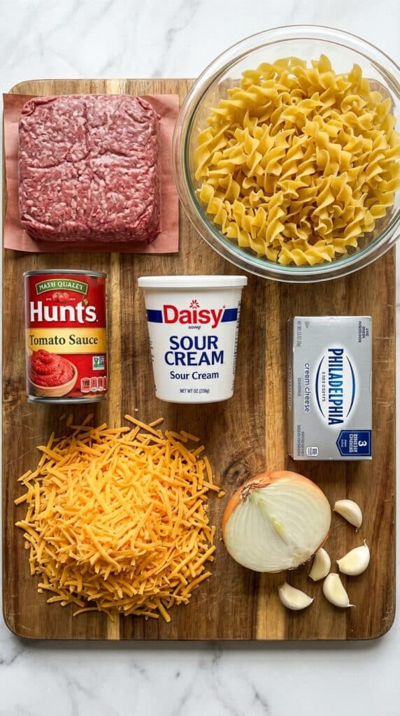 A flat lay showing ground beef, dry egg noodles, tomato sauce, sour cream, cream cheese, cheddar cheese, and an onion on a wooden board.