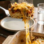 A close-up of a spatula lifting a hearty square of ground beef and noodle casserole, creating a massive cheese pull from the pan.