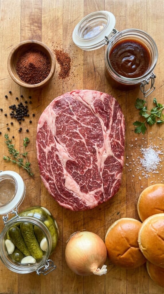 A flat lay showing a raw beef chuck roast, spice rub, BBQ sauce, a jar of pickles, onions, and brioche buns on a butcher block.