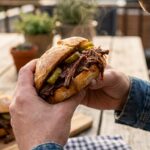 A close-up of two hands holding a juicy, messy pulled beef sandwich with pickles, about to take a bite.