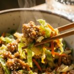 A close-up of wooden chopsticks lifting a savory bite of pork, shredded cabbage, and carrots from an egg roll in a bowl.