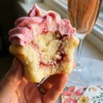 A close-up of a hand holding a half-eaten raspberry swirl cupcake, showing the fluffy interior and marbled pink frosting.