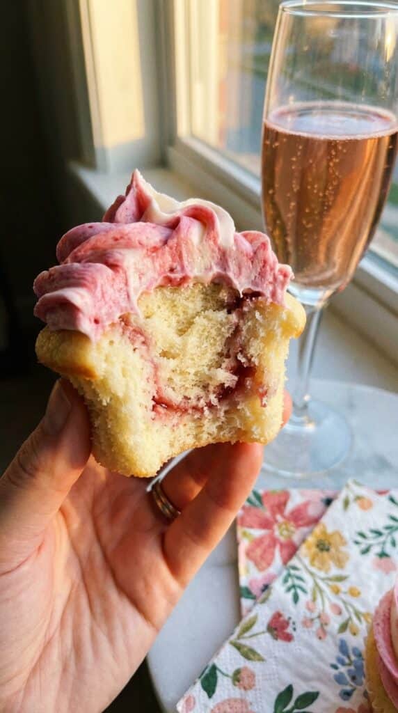 A close-up of a hand holding a half-eaten raspberry swirl cupcake, showing the fluffy interior and marbled pink frosting.