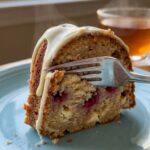 A close-up of a slice of raspberry white chocolate Bundt cake on a plate, with a fork cutting into the moist crumb showing baked berries and chocolate.