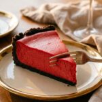 A close-up of a slice of red velvet cheesecake on a plate with a fork cutting into the tip, showing the smooth, dense red filling.
