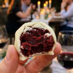 A close-up of a hand holding a white chocolate truffle with a bite taken out, revealing a dense, fudgy red velvet center.