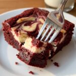 A close-up of a fork breaking into a fudgy red velvet cheesecake brownie, showing the dense texture, with a glass of milk in the background.