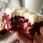 A close-up of a hand holding a red velvet cookie dough bar with a bite taken out, showing the dense red dough and white chocolate chips inside.