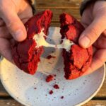 Two hands breaking apart a warm red velvet cookie, revealing a soft, gooey red center and melted white chocolate chips.