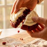 A close-up of hands breaking a frosted red velvet cookie in half, showing a soft, chewy red center and thick white frosting.