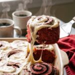 A close-up of a spatula lifting a bright red velvet cinnamon roll from a pan, showing a stretch of melted white cream cheese frosting.