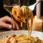 A close-up of a silver fork twirling glossy linguine and a shrimp over a pasta bowl, with a hand holding a wine glass in the background.
