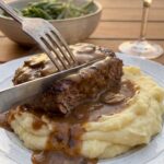 A close-up of a knife and fork cutting into a Salisbury steak covered in mushroom gravy, served over a bed of mashed potatoes.