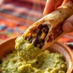A close-up of a hand dipping a half-eaten crispy vegan taquito into a bowl of fresh guacamole.