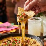 A close-up of a tortilla chip scooping up thick, melted sausage cheese dip with a massive cheese pull in a party setting.