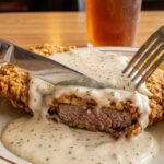 A close-up action shot of a knife and fork cutting into a crispy chicken fried steak smothered in pepper gravy, showing the tender beef inside.