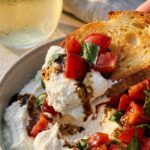 A close-up of a piece of toasted bread scooping up whipped ricotta, diced tomatoes, basil, and balsamic drizzle from a bowl.