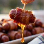A close-up of a hand holding a bacon-wrapped cocktail sausage by a toothpick, with a thick drop of caramelized brown sugar dripping from it.