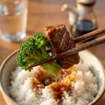 A close-up of wooden chopsticks holding a piece of saucy beef and broccoli over a bowl of white rice.