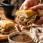 A close-up of hands dunking a cheesy beef sandwich into a bowl of dark, steaming au jus.