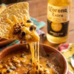 A close-up of a tortilla chip scooping up thick, cheesy nacho soup with a string of melted cheese stretching from the bowl.